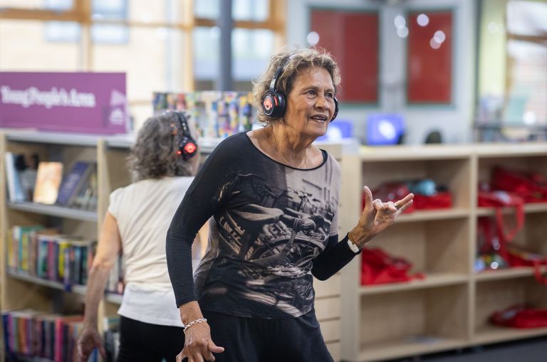 Two senior women dance with headphones on, enjoying music in a library setting.